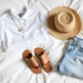 Sunlit flatlay of summer fashion essentials including a white linen shirt, denim shorts, straw hat, sandals, and gold jewelry, arranged on white bedding with soft morning light.