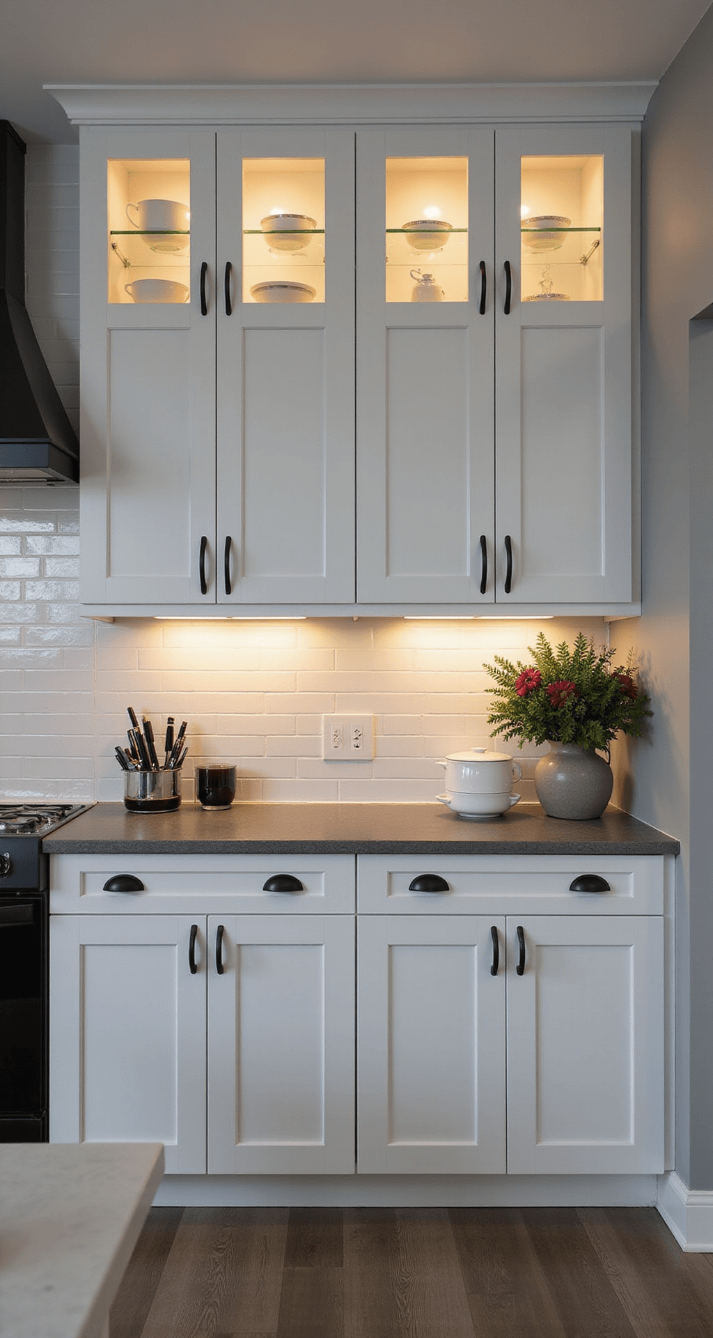 Intimate view of a transitional kitchen at dusk, featuring modern white cabinets with integrated handles, moody accent lighting, glass-front upper cabinets with curated dishware, matte black fixtures, quartz countertops with waterfall edges, and minimalist decor including a dramatic floral arrangement, in a color palette of pure white, matte black, cool grey, and greenery.