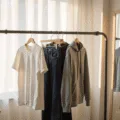 A sunlit minimalist dorm room showcasing a sleek clothing rack with white t-shirts, dark wash jeans, and a gray hoodie, illuminated by golden hour light filtering through white curtains. A full-length mirror reflects the organized wardrobe, and a stack of textbooks is neatly placed nearby, all in soft, warm tones.