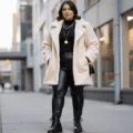 A confident plus-size model poses in a cream teddy coat, black leather leggings, and combat boots during golden hour, with layered gold necklaces and a fitted black turtleneck bodysuit, against a softly blurred urban background.