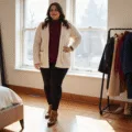 A plus-size model in a burgundy turtleneck and cream chunky knit cardigan poses confidently in a sunlit modern bedroom, complemented by black fleece-lined leggings and cognac leather boots, with winter coats in jewel tones on display nearby.