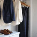 Bright walk-in closet with a navy blazer, cream silk blouse, and tailored black trousers on wooden hangers, polished leather loafers on a shelf, minimal gold jewelry on a marble tray, illuminated by soft morning light.