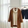 A minimalist bedroom with a white clothing rack displaying a winter capsule wardrobe, including cream chunky knit sweaters, dark jeans, cognac leather ankle boots, and a camel wool coat, complemented by beige cashmere accessories on a marble surface, all bathed in natural light.