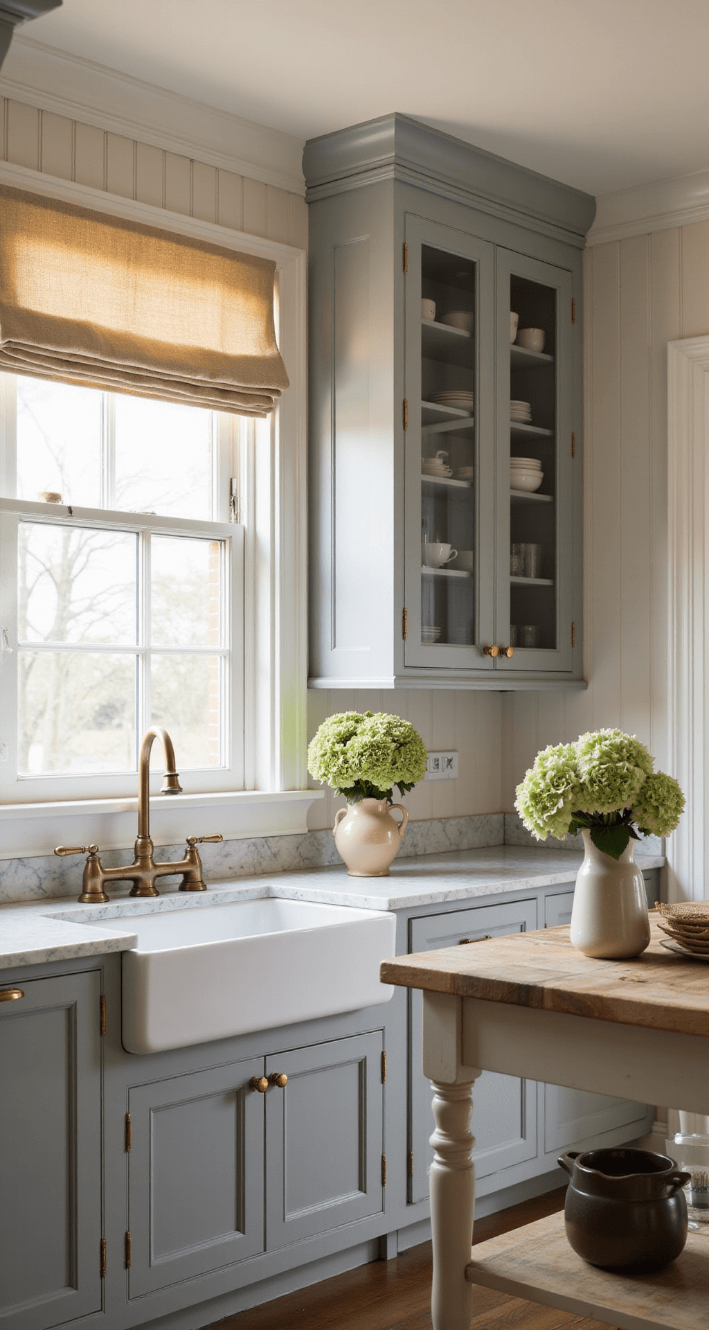 An elegant BODBYN kitchen with soft gray raised-panel cabinets and brass hardware at golden hour, showcasing glass-front upper cabinets filled with vintage dishware, marble countertops, and a farmhouse sink. A butcher block island with turned legs complements the space, adorned with fresh hydrangeas in a ceramic vase. Natural linen window treatments filter warm light, capturing a timeless blend of traditional and modern styles.