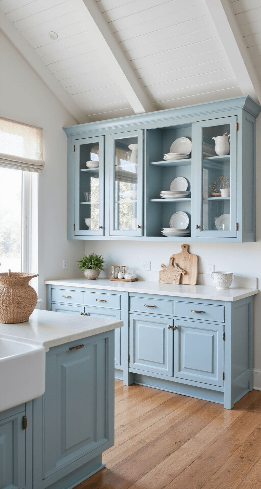 A sunlit coastal kitchen featuring pale blue cabinets with seeded glass doors, displaying white ironstone and sea glass, under a vaulted white-planked ceiling, with a beachy color palette of soft blues, whites, and sandy beige.