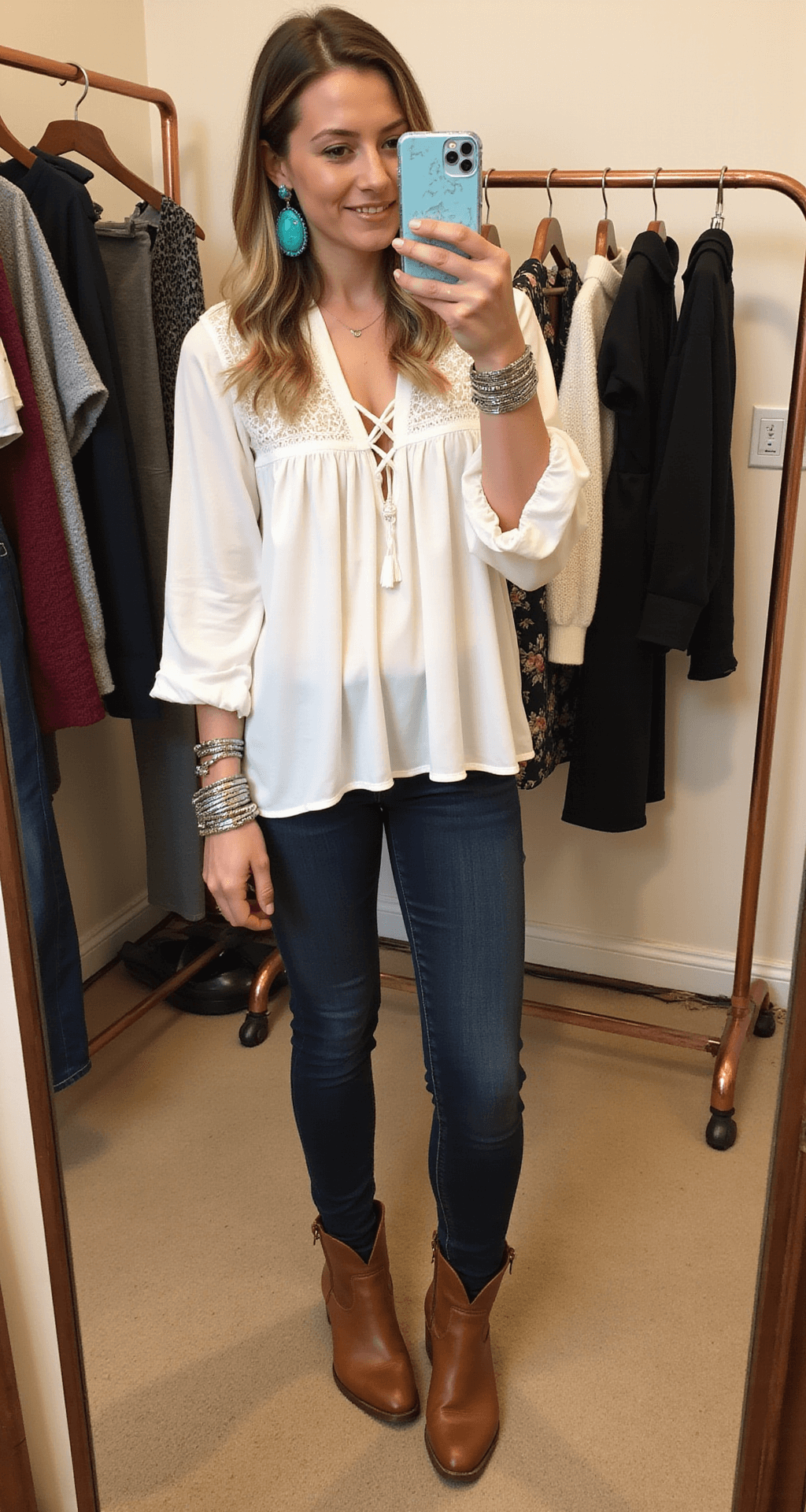 A woman in a boutique dressing room tries on a flowy white peasant blouse with dark skinny jeans and cognac leather boots, while turquoise earrings and silver bangles reflect in the mirror. Elegant outfits hang on a copper rack, illuminated by warm interior lighting.