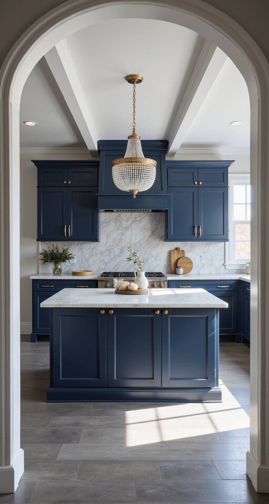 Transitional navy kitchen featuring coffered ceiling, bright indirect sunlight, navy inset cabinets with brass and chrome accents, white marble counters with dramatic veining, geometric grey and white tile backsplash, crystal chandelier over the island, and grey wood-look tile flooring, viewed from the entrance.
