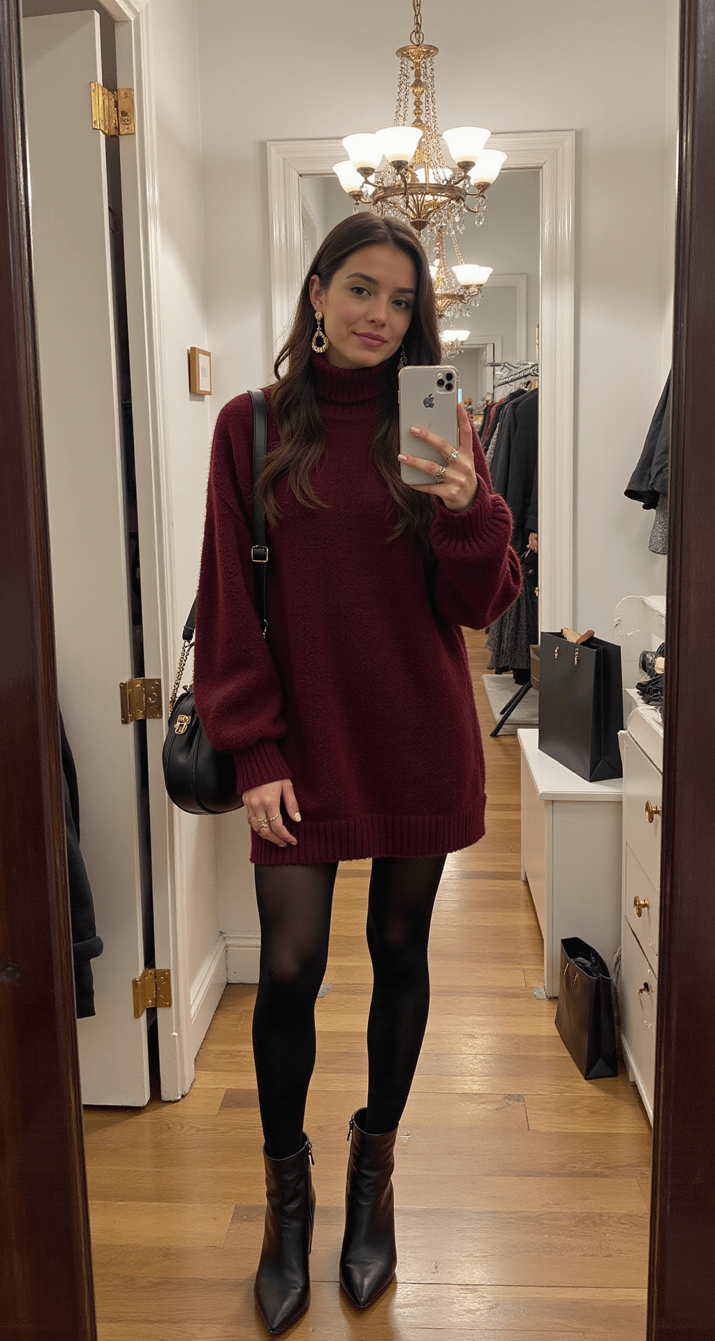 A model in a burgundy turtleneck sweater dress and black opaque tights is reflected in a mirror inside a boutique dressing room with vintage chandelier lighting. She wears pointed-toe ankle boots, statement gold earrings, and multiple delicate rings, with shopping bags visible in the background, highlighting the outfit's proportions and styling details.