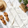 A bright and airy flatlay featuring a white cotton sundress, a woven straw sunhat with shell details, layered gold necklaces, leather sandals, and a rattan clutch on white linen, accented by tropical flowers and a coconut drink.