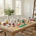 Cozy craft room with white shiplap walls and oak floors, featuring a large farmhouse table adorned with glass ornaments and vibrant glitters in gold, silver, red, and emerald green. Natural light illuminates the scene, highlighting hands at work and sparkling finished ornaments on a rustic shelf.
