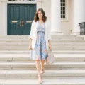 An elegant woman in a powder blue floral dress and cream cardigan ascends sunlit stone steps, wearing nude heels and carrying a beige handbag, surrounded by architectural details in soft natural lighting.