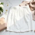 Bright editorial flat lay of a white pleated midi skirt, gold jewelry, beige cashmere sweater, and nude leather sandals on a marble desk, accented by a designer handbag, white peonies, and silk scarf, illuminated by natural sunlight.