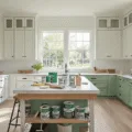 Photorealistic wide-angle view of a modern kitchen renovation in progress, showcasing cabinet doors in various transformation stages from original wood to a sage green finish, with organized painting supplies and soft morning light filtering through large windows.