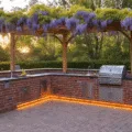Photorealistic image of a luxurious L-shaped brick BBQ station at golden hour, featuring custom herringbone-patterned red clay bricks, polished granite countertops, and a cedar pergola draped in wisteria, with integrated LED lighting and a curved seating area with navy cushions.