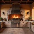 A luxurious Mediterranean outdoor kitchen featuring a stainless steel grill and wood-fired pizza oven under stone archways, adorned with terracotta tiles and a pergola draped in bougainvillea, illuminated by warm sunset light.