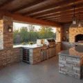 Photorealistic wide-angle shot of a luxurious L-shaped outdoor kitchen during golden hour, featuring a stainless steel grill, waterfall granite countertops, stone veneer walls, and a cedar wood pergola, with modern pendant lighting, a built-in wine fridge, and a brick pizza oven, surrounded by fresh herbs in terracotta planters.