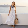 Model in a white linen maxi dress on a Santorini balcony at golden hour, with the Aegean Sea in the background, styled with leather sandals, a straw tote, gold jewelry, and a blue silk scarf.