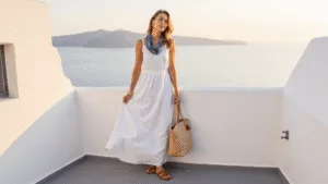 What to Wear in Greece: Your Ultimate Style Guide (That Actually Works!) Model in a white linen maxi dress on a Santorini balcony at golden hour, with the Aegean Sea in the background, styled with leather sandals, a straw tote, gold jewelry, and a blue silk scarf.