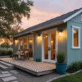 Ultrawide cinematic photograph of a modern garden shed in soft sage green with white trim, featuring floor-to-ceiling windows, French doors, a minimalist floating deck, and surrounded by ornamental grasses and lavender, all captured at golden hour in photorealistic detail.