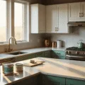 A sunlit modern kitchen showcasing a transformation of a white Shaker-style cabinet with a sage green island, featuring an organized array of painting supplies in the foreground, and a partially-painted cabinet door demonstrating before/after contrast, all captured with dramatic lighting and depth of field.