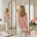 A woman in a blush cashmere sweater admires her reflection in a triple mirror within a minimalist dressing room adorned with white marble and brass fixtures, complemented by pink roses and sunlight streaming through the space.