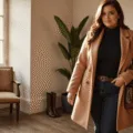 Plus-size model in a luxurious camel wool-blend coat, black turtleneck, and dark-wash jeans in a sunlit modern dressing room, with warm golden hour lighting.
