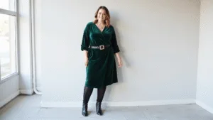Plus Size Dressy Winter Outfits: Your Complete Style for 2024 Plus-size model in an emerald velvet wrap dress styled with black fleece-lined tights, knee-high leather boots, a gold statement belt, and delicate gold jewelry against a minimalist backdrop, showcasing an elegant winter formal look.