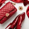 A luxurious flat lay of rich red fashion accessories including a quilted leather handbag, patent leather boots, a flowing silk scarf, a velvet hair bow, and bold red lipstick, accented with delicate gold jewelry, arranged on a white background with soft lighting.