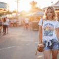A young woman in high-waisted denim shorts and a vintage graphic tee stands at a fairground entrance during golden hour, with a light denim jacket tied around her waist, a small leather crossbody bag, and white sneakers. Colorful carnival lights and vibrant booths create a lively background.