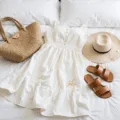 A bright flatlay of summer fashion essentials on white bedding, including a flowy white sundress, neutral linen pieces, leather sandals, straw accessories, and delicate gold jewelry, all illuminated by natural light.