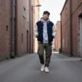 A confident young person wearing an oversized navy and cream varsity jacket over a grey hoodie, olive cargo pants, and cream sneakers, walking against a weathered brick wall during golden hour, with dramatic shadows and a muted urban color palette.