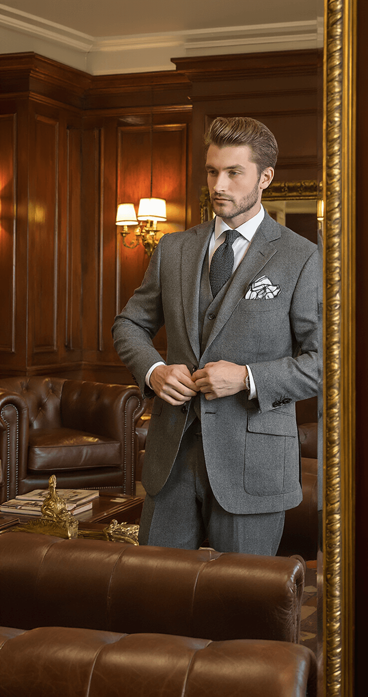 Male model in a grey flannel suit jacket adjusts his pocket square, reflected in an antique mirror within a private men's club lounge. The setting features mahogany paneled walls, leather club chairs, and brass fixtures, all illuminated by warm ambient lighting from vintage sconces, enhancing the traditional atmosphere.