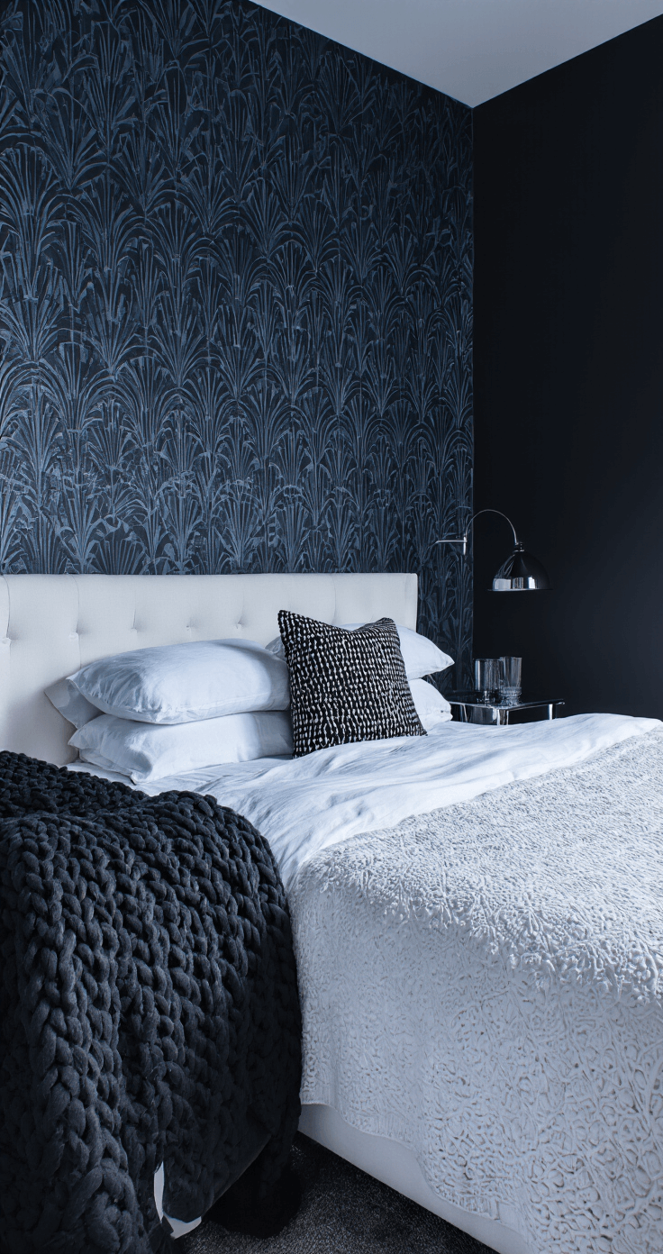 A cozy 12x12ft bedroom at blue hour, featuring black geometric art deco wallpaper on the focal wall, a white upholstered bed with textured bedding and a chunky black knit throw, and chrome and glass side tables reflecting deep twilight, with dramatic side lighting enhancing the wall textures.