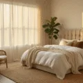 A serene 14x16ft bedroom filled with warm golden hour light, featuring a plush king-size bed with layered bedding, a vintage rattan reading chair, and a fiddle leaf fig, all set against warm greige walls adorned with black and white photographs.
