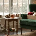 A cozy craft corner adorned for Christmas, featuring a plush green armchair, a distressed oak side table with a mug of cocoa, a vintage craft box filled with festive supplies, and soft sunlight streaming through a bay window, all highlighted by twinkling fairy lights and holiday decorations.