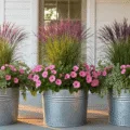 A golden hour photograph of a rustic farmhouse porch with galvanized metal planters showcasing tall purple fountain grass, soft pink petunias, and cascading silvery dichondra, set against whitewashed wood and vintage brick, with dramatic shadows and warm atmospheric lighting.