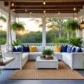 Photorealistic image of a luxurious coastal retreat's wraparound porch at golden hour, featuring a white wicker sectional, macramé swing chair, blue ceramic planters with ferns, and market lights, framed by tropical palms and white colonial columns.