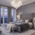 A luxurious master bedroom suite with a tufted grey velvet king bed, crystal chandelier, marble nightstands, and floor-to-ceiling windows, all set in a twilight ambiance.