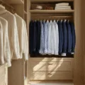 A luxurious walk-in closet with oak built-ins displaying neatly arranged cream cashmere sweaters, white button-downs, and navy blazers, illuminated by soft afternoon light with leather accessories and gold hardware accents.