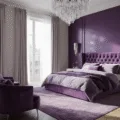 Photorealistic interior of a luxurious purple master bedroom featuring a deep purple accent wall, plush king-size bed with lavender velvet headboard, layered bedding, crystal chandelier, and elegant decor, bathed in soft golden hour light.