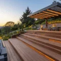 A luxurious multi-level deck in a terraced garden during sunset, featuring rich walnut composite decking, modern glass railings, sleek outdoor furniture, and a contemporary pergola, all bathed in warm golden light.