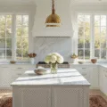Photorealistic image of a bright, airy kitchen with white shaker cabinets, a Carrara marble island, brass hardware, stainless steel appliances, and natural morning light streaming through floor-to-ceiling windows, featuring decorative elements like a vintage runner and white hydrangeas.