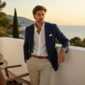 A male model in a white linen shirt and beige chinos leans against a whitewashed railing on a coastal terrace at golden hour, with a navy blazer draped on a chair, brown loafers, and a Panama hat; the Mediterranean Sea and cypress trees are visible in the warm evening light.