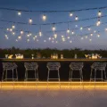 A luxury outdoor bar at twilight featuring a matte black aluminum counter, modern chrome barstools, ambient string lights, tropical plants, and crystal-clear cocktail glasses, all presented in a photorealistic architectural style.