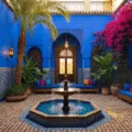 A hyperrealistic wide-angle view of a serene Moroccan courtyard garden at golden hour, featuring Majorelle blue walls, intricate zellige tiles, a central brass fountain, dappled shadows from date palms, vibrant magenta bougainvillea, brass lanterns casting decorative shadows, a plush seating area with jewel-toned cushions, and lush greenery in blue glazed pottery.