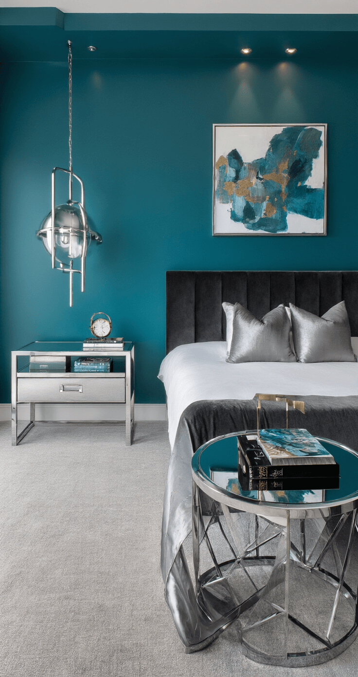A contemporary bedroom featuring a deep peacock teal statement wall, dusk lighting from hidden LED coves, a low-profile charcoal velvet bed with silver silk pillows, chrome and glass side tables, and abstract art in blues and metallics, shot from an elevated angle.