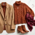 Overhead flatlay of fall fashion items including a camel wool blazer, rust chunky knit sweater, brown leather ankle boots, gold jewelry, and a burgundy silk scarf arranged on a white marble surface, with soft natural light enhancing the textures.