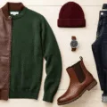 Flat lay of men's fall fashion essentials on a light wooden surface, featuring a brown leather bomber jacket, forest green merino wool sweater, dark wash denim jeans, brown leather Chelsea boots, burgundy knit beanie, and a silver watch, all arranged with natural lighting and shadows, evoking a sophisticated autumn vibe.